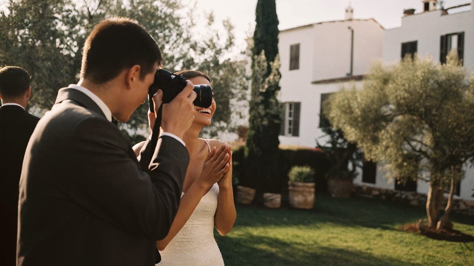 Un amigo de los novios fotografía a la novia riendo con una réflex en el jardín