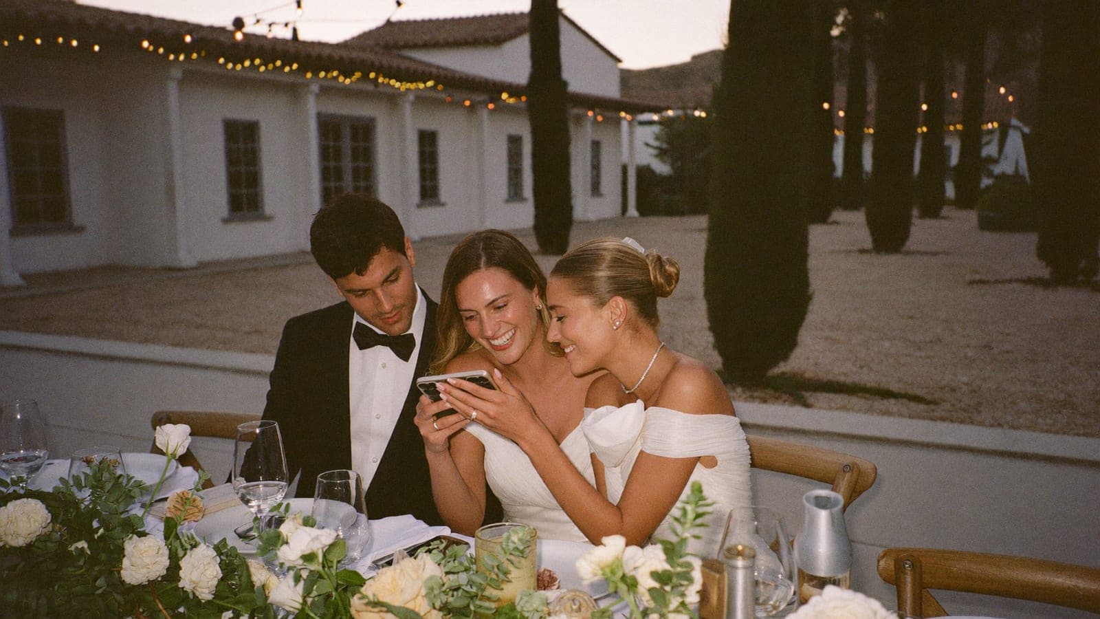 Los novios y una invitada se ríen mirando fotos en un solo móvil en la mesa del banquete