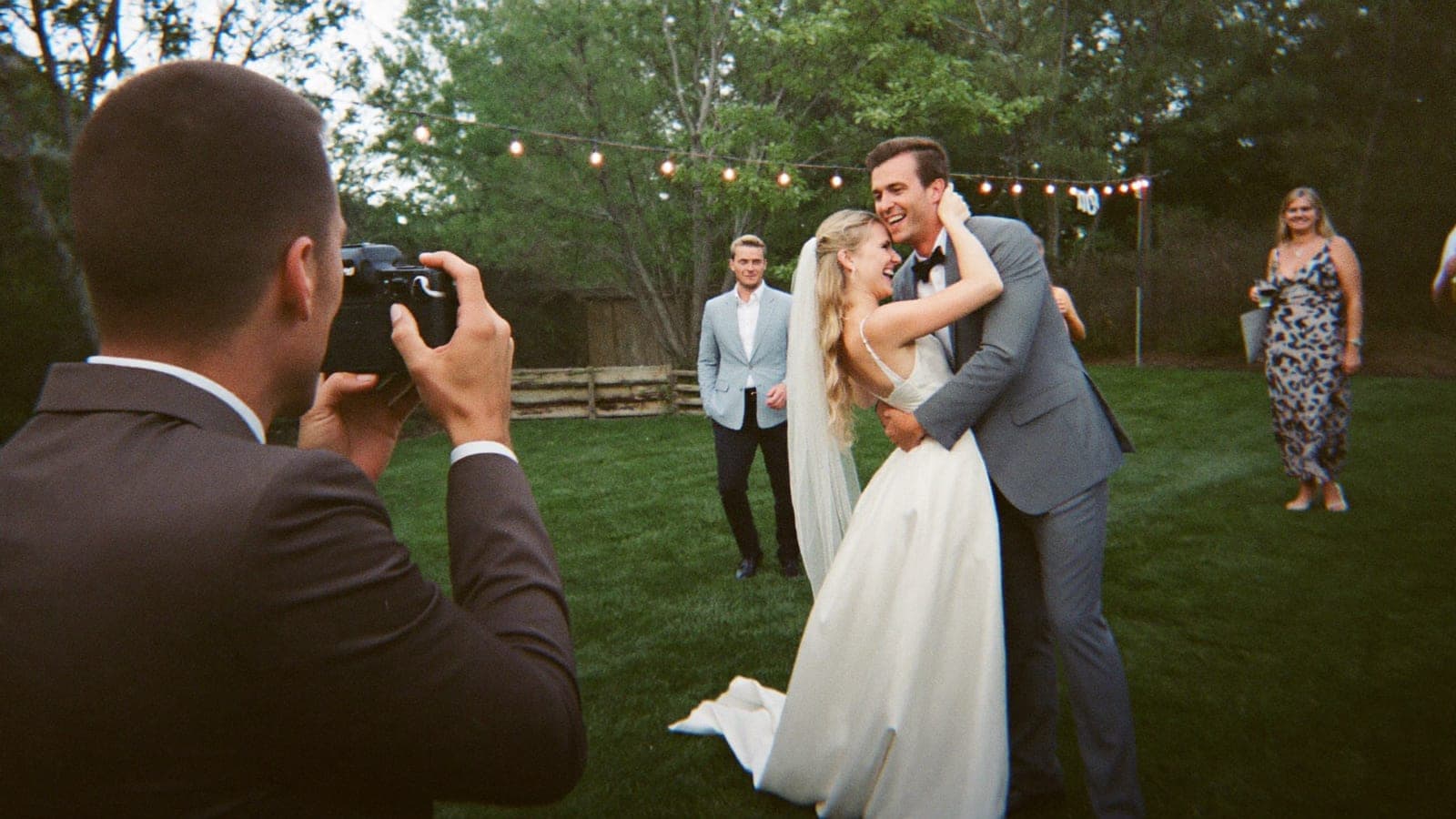 A friend of the couple photographing the laughing newlyweds with a DSLR at a backyard wedding