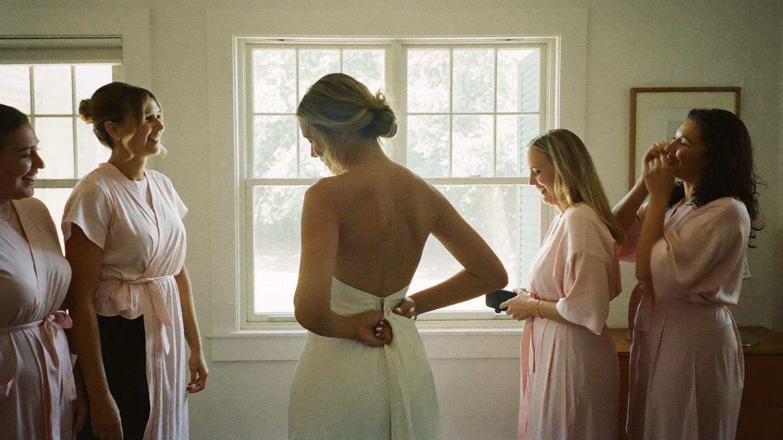 The bride's mother does up the buttons of the wedding dress by the window