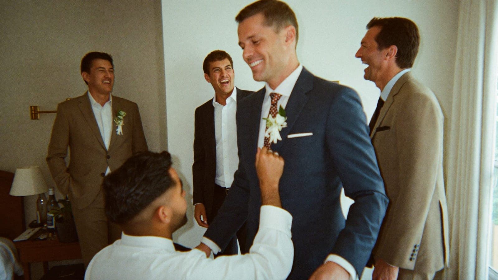 The groom laughing with his groomsmen as one pins on his boutonniere on the wedding morning