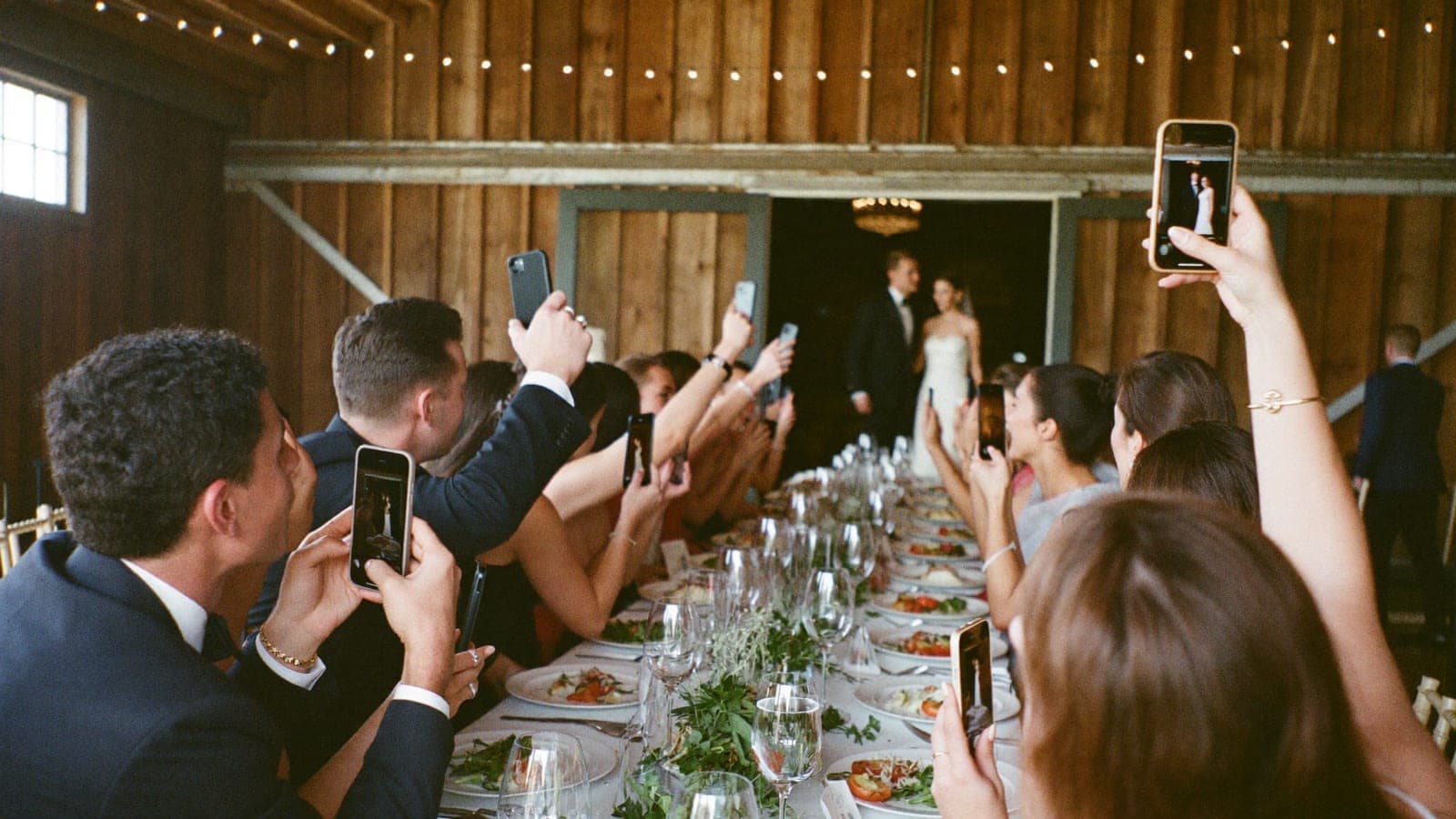 Guests at the dinner table raising their phones as the couple walks in